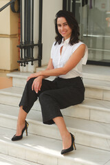 An elegant attractive young woman in a white blouse and black trousers sits on the steps and looks at the camera smiling.