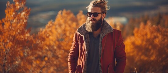 A male model with a hipster vibe of Caucasian descent posing outdoors in the midst of nature The landscape showcases vibrant colors with trees and a rural road lined with orange and red lea - obrazy, fototapety, plakaty