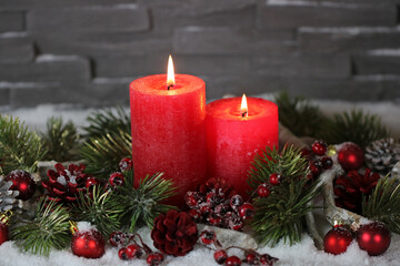 Two red burning candles in the snow with Christmas decorations and fir branches.