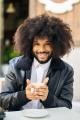 a smiling young man drinking a hot coffee on the terrace of a coffee shop