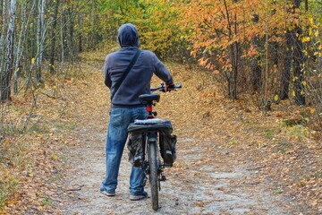 a tourist in a gray fashionable raglan with a hood on his head and blue stylish jeans goes with a sports mountain black modern new bicycle in the forest  during  - obrazy, fototapety, plakaty