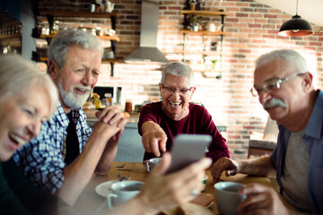 Group of senior friends using a smartphone while having breakfast together in the kitchen - obrazy, fototapety, plakaty