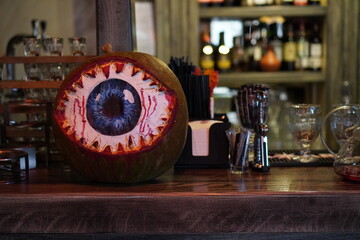 a large pumpkin with a painted eye on a wooden bar counter table against the background of shelves with a mirror and bottles of drinks at a Halloween party