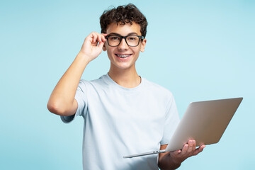 Smart smiling teenager boy wearing eyeglasses and casual t shirt looking at camera holding laptop