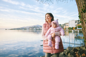 Outdoor portrait of happy young mother with adorable baby girl enjoying nice view of winter lake...
