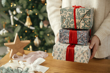 Woman holding stylish christmas gifts with red ribbon at wooden table with festive decorations on background of decorated tree in scandinavian room. Merry Christmas and happy holidays!