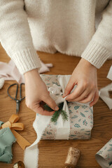 Woman wrapping stylish christmas gift with ribbon and fir branch on wooden table with festive...