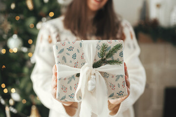 Merry Christmas and happy holidays! Woman holding stylish christmas gift with ribbon and fir branch  close up on background of modern decorated tree in scandinavian room.