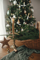 Merry Christmas and Happy holidays! Stylish rustic basket with fir branches, vintage candle and wooden star on table against festive decorated christmas tree in scandinavian room.