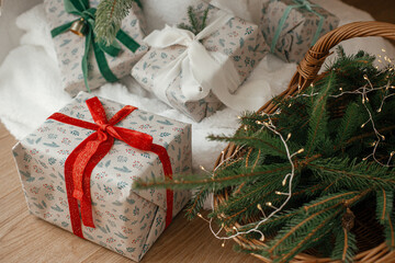 Stylish wrapped christmas gifts and rustic basket with fir branches under festive decorated christmas tree in scandinavian room. Merry Christmas and Happy holidays!