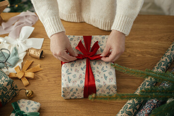 Woman wrapping stylish christmas gift with red ribbon on wooden table with festive decorations in...