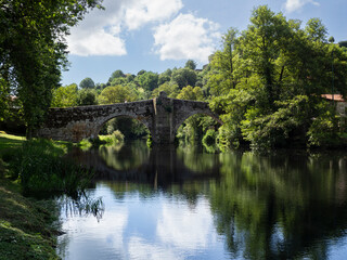 Puente antiguo de piedra con dos arcos sobre el río de Allaritz, reflejado en el agua junto al cielo y los árboles verdes alrededor de la orilla, viajando por Galicia en España, verano de 2021