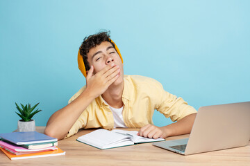 Attractive smiling teenager boy wearing casual clothes yawning sitting at desk using laptop - obrazy, fototapety, plakaty