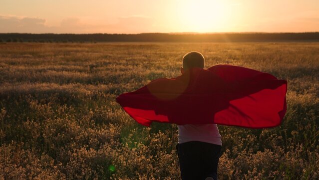 Child superhero in red cape, nature. Active Boy, child plays superhero, sun. Happy run of boy child in red raincoat, sunset, park. Kid runs across green field with flowers, childhood dream kid to fly