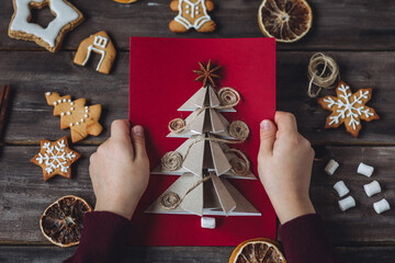 Idea for easy Christmas do it yourself crafts for kids to make. Child's hands with handmade postcards with fir tree and toy from recycled cardboard paper, thread, buttons. Wooden background, flat lay