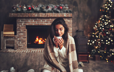 Jeune femme brune assise face caméra, portant un pull blanc et emmitouflée dans un plaid, qui sirote un chocolat chaud devant une cheminée avec un feu, arbre de noël et décorations derrière elle. - obrazy, fototapety, plakaty