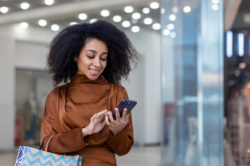 Young beautiful woman shopper inside supermarket store using app on phone, holding colorful shopping gift bags and smartphone, smiling happily.