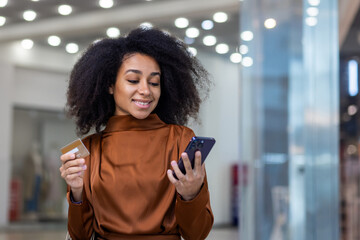 Young beautiful woman inside a supermarket large clothing store in a shopping mall using a smartphone app online shopping, an African American woman smiling contentedly holding a bank credit card.