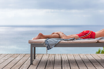 Les jambes d'un  homme en maillot de bain sur transat en terrasse, serviette pend du transat, mer...