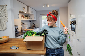 Smiling woman eating fresh carrot from her healthy food delivery box. Box with vegetables and phone...