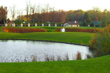 Obraz premium Picturesque landscape featuring a serene pond with a fountain, lush green grass, autumnal trees and a charming cottage in the background, ideal for tranquil and scenic photography.