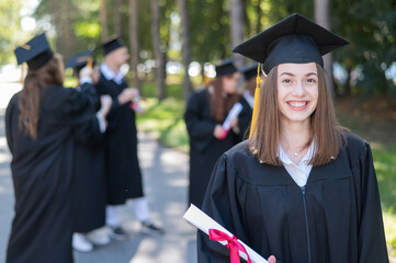 Group of happy students in graduation gowns outdoors. A young girl with a diploma in her hands in...