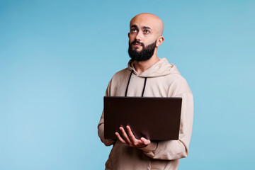 Thoughtful arab man holding laptop and looking upwards with pensive expression. Young person in doubt programming software application while standing with portable computer