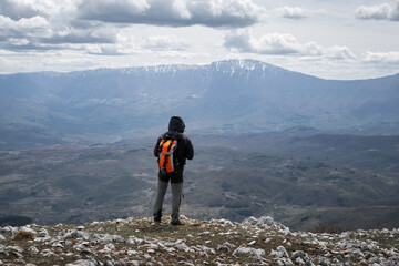 Mountain hiker with orange backpack standing at the edge of the cliff and looking at Suva planina (Dry mountain) and snow covered Trem summit - 671280773
