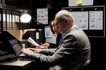 Professional elderly businessman manager and asian assistant checking administrative analytical data in file cabinet office. Accounting team in bureaucratic office filled with invoice folders - obrazy, fototapety, plakaty