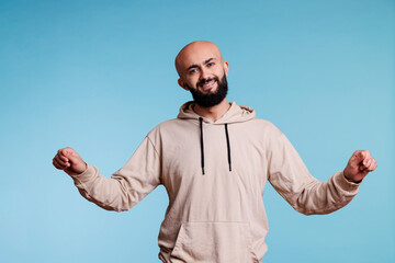 Fototapeta premium Smiling arab man pretending to hold banner in two hands and looking at camera with cheerful facial expression. Young adult joyful person advertising product studio portrait