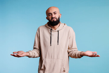 Fototapeta premium Arab man with spread arms and puzzled expression showing do not know gesture portrait. Young bald beared person looking at camera with confused emotions while shrugging shoulders