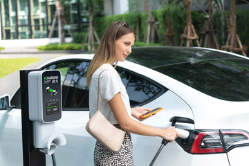 Young woman travel with EV electric car charging in green sustainable city outdoor garden in...
