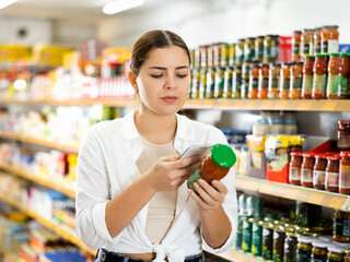 in supermarket, young woman thoughtfully chooses tomato sauce.Woman scans qr code from sauce label