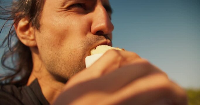 Close-up shot of the face of a happy brunette guy with stubble who eats a hot dog. A cheerful guy eats a hot dog and smiles during his day off