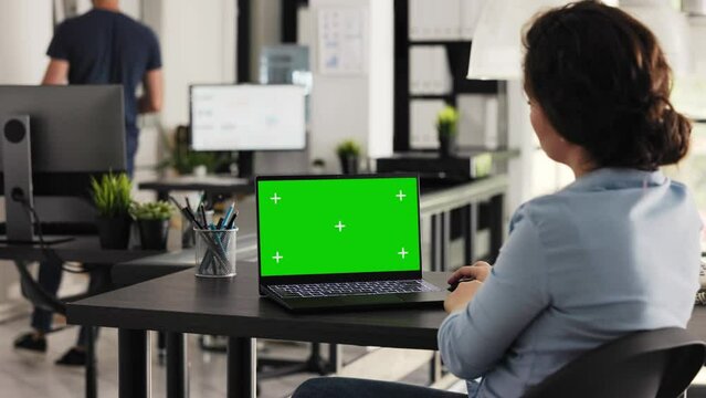 Businesswoman works on laptop with greenscreen, looking at blank chromakey display in coworking space. Young adult examining pc screen with isolated mockup template, e commerce tasks. Handheld shot.