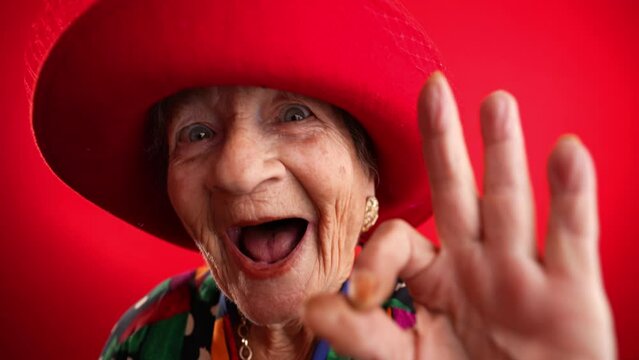 Happy fisheye portrait caricature of funny elderly woman with red hat giving OK sign gesture with hand isolated on red background.