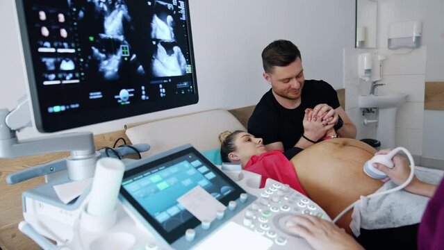 Woman on the last stages of pregnancy is checked on the ultrasonic equipment. Caring man supporting his husband.