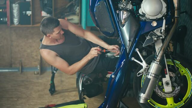 Vertical video of a male master Motor mechanic with a short haircut in a gray T-shirt repairing his blue and yellow bike and dismantling it into pieces in his workshop garage
