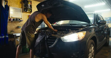 A male master mechanic with a short haircut in a gray T-shirt and green shorts repairs the hood of a car with the headlights on and a slight haze in his garage workshop