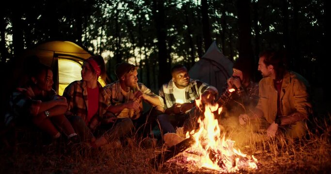 A group of scouts during a rest stop tell each other scary stories near a fire in the night forest with tents in the background