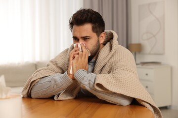 Sick man wrapped in blanket with tissue blowing nose at wooden table indoors. Cold symptoms