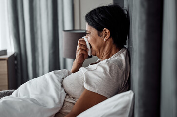 Sick, tissue and elderly woman in bed blowing nose, flu or viral infection in her home. Allergies, bacteria and senior female sneezing, coughing or sore throat in a bedroom with virus or covid