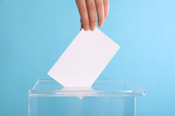 Woman putting her vote into ballot box on light blue background, closeup