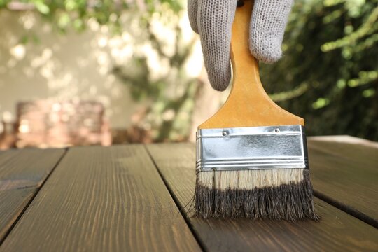 Man applying wood stain onto wooden surface outdoors, closeup. Space for text