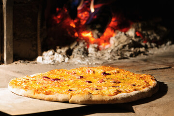 A classic whole italian pizza n the door of a clay oven with wood fire behind
