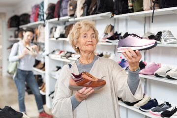 Fototapeta na wymiar Mature european woman who came to a shoe store for shopping, chooses sports sneakers, standing near the shelves with the goods