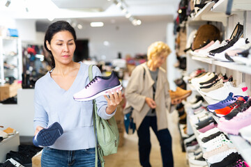 Asian woman selecting new sport shoes while standing in salesroom of shoeshop. Elderly European...