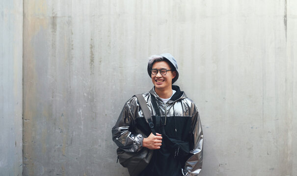 Fashion, student and gen z asian male happy, confident and . with cool clothes on wall background, smile and chilling. Travel, fashionable and Japanese guy outdoors casual, contemporary and stylish