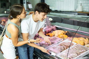 Attentive young man and woman purchasers choosing sausages from closed-type showcase in butcher shop