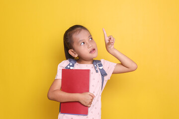 Adorable Asian girl student holding book and pointing up to empty space isolated on yellow...